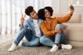 Sitting comfortably on the floor, a delighted young couple enjoys a light-hearted moment taking a selfie