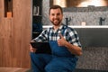 Sitting on the floor with notepad in hands. Plumber in blue uniform is at work in the bathroom