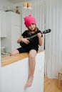 Sitting on a high table little girl in a pink hat playing on a guitalele.