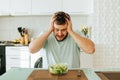 Sitting in the kitchen, a young man looks with horror at a green salad that he should eat.