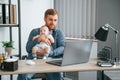 Sitting by the table with laptop. Father with toddler is indoors in the office