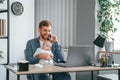 Sitting by the table with laptop. Father with toddler is indoors in the office