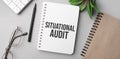 situational audit is written in a white notebook with calculator, craft colored notepad, plant, black marker and glasses