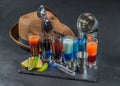 six different colored shot drinks, lined up on a black stone plate, ice cubes in shaker and ice tongs, lemon and lime, brown Royalty Free Stock Photo