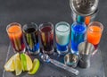 six different colored shot drinks, lined up on a black stone plate, ice cubes in shaker and ice tongs, lemon and lime Royalty Free Stock Photo