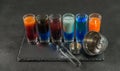 six different colored shot drinks, lined up on a black stone plate, ice cubes in shaker and ice tongs Royalty Free Stock Photo