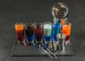 six different colored shot drinks, lined up on a black stone plate, ice cubes in shaker and ice tongs Royalty Free Stock Photo