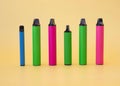 Six electronic vape cigarettes of different colors and sizes stand on a yellow background