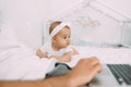 A six-month-old baby lies on a white bed and watches dad study while sitting next to a laptop. Royalty Free Stock Photo