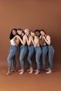 Six multiracial females in casuals hugging together on brown background in studio. Group of women of different ages standing