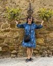 Sixty-five year-old female Korean tourist posing beneath flower baskets on a street in Pienza, Italy.