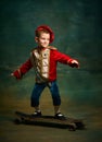 Smiling little boy dressed up as medieval little prince and pageboy skateboarding over dark vintage style background