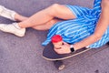 Skater woman in dress is sitting on longboard with paper cup of coffee. Close up view on hand and board.