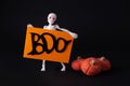 Skeleton holding a poster with the words boo and pumpkins from marshmallow on a black background, halloween concept