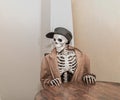 Skeleton wearing brown leather jacket with leather hat, sitting behind the table