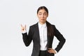 Skeptical and awkward young asian businesswoman, saleswoman in black suit shaping something small and looking