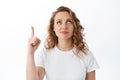 Skeptical and doubtful blond woman grimacing, pointing and looking up unsure, standing against white background