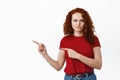 Skeptical and upset redhead girl grimacing, frowning and smirking while pointing fingers left at bad company logo