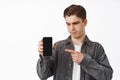 Skeptical young man looks doubtful, pointing and showing smartphone screen, interface or app on mobile phone, grimacing Royalty Free Stock Photo