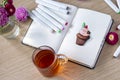 Sketching cupcake in a notebook. All placed on a wooden table with a transparent cup of tea, notebook, markers, and flowers.Hobby