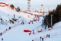 A ski slope for beginners with a lot of people on it and a fence, a chapel and trees on top of the slope. Russia - January 30, Royalty Free Stock Photo