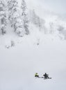 A skier and a snowboarder talking reclining on a steep mountain slope near a forest in a snowstorm