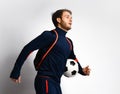 Skilled male soccer player in blue tracksuit and backpack is running, holding a ball, posing sideways isolated on white. Close up