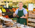 Skilled mature male supermarket worker in apron selling various olives and pickles in grocery supermarket