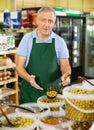 Positive old male merchandiser in apron picking up olives with scoop from plastic container in grocery store