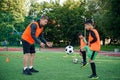 Skillful teen player in football uniform working out the kicking ball together with his experienced coach on sport field
