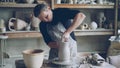 Skillful young potter is molding ceramic vase from clay on turning throwing wheel while working in potter& x27;s workshop