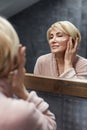 Skincare Routine. Mature Woman Uses Cosmetic Cream On Face Skin In Front Of The Mirror Reflection.