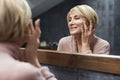 Skincare Routine. Mature Woman Uses Cosmetic Cream On Face Skin In Front Of The Mirror Reflection.