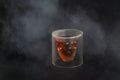 Skull shaped glass of brown alcohol drink and white smoke or steam on black background.