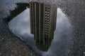 A skyscraper's reflection is vividly captured in a puddle on a textured road surface, under a cloudy sky. Royalty Free Stock Photo