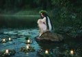 Slavic girl sits on stone on shore lake. Nymph fantasy woman hugs knees. Long black hair. Wreaths of grass, flowers Royalty Free Stock Photo