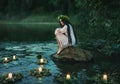 Slavic girl sits on stone on shore lake. Nymph fantasy woman hugs knees. Long black hair. Wreaths of grass, flowers