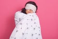 Sleep equipment concept. Portrait of brunette woman wrapped white blanket and wearing sleeping eye mask. Studio shot of young