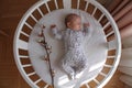 A sleeping baby girl in a bodysuit lies in a white round crib with a cotton flower