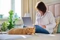 Sleeping ginger cat at home on bed, woman using laptop on background Royalty Free Stock Photo