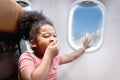 Sleepy curly hair African girl child passenger sitting in comfortable seat next to window inside airplane, kid yawning and Royalty Free Stock Photo