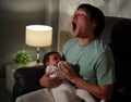 Sleepy father yawning and feeding milk bottle to infant baby on sofa in the living room at night