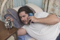 A sleepy and groggy man lying in bed checks the time on his alarm clock while talking to a client. Freelancer taking up rush work