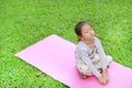 Sleepy little Asian kid girl sitting on pink mattress in green grass lawn. Closed eyes child outdoor