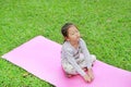 Sleepy little Asian kid girl sitting on pink mattress in green grass lawn. Closed eyes child outdoor