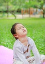 Sleepy little Asian kid girl sitting on pink mattress in green grass lawn. Closed eyes child outdoor
