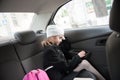 Sleepy little girl in autumn black coat and white hat on trip to school in morning sitting on back seat of safe car with pink Royalty Free Stock Photo