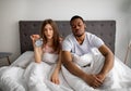 Sleepy multiracial couple sitting on bed with alarm clock, cannot open their eyes, too tired to get up in morning Royalty Free Stock Photo