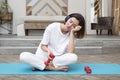 Sleepy woman doing sport and listening to music with headphones on fitness mat at home with dumbbells