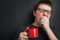 Sleepy yawning man in eyeglasses with red cup of tea or coffee has uncombed hair in underwear on black background, morning refresh